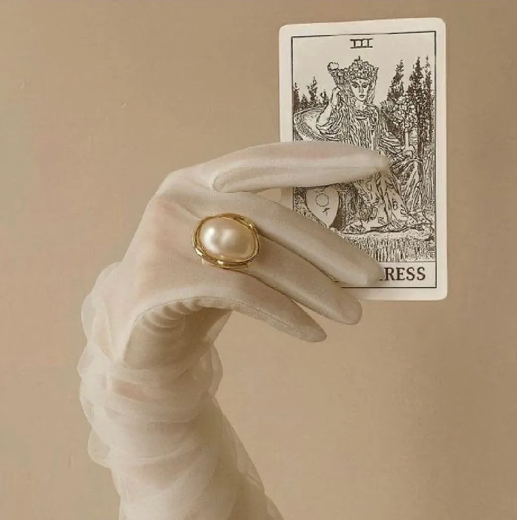 Mão com luva branca e anel de pérola segurando uma carta de tarô.
