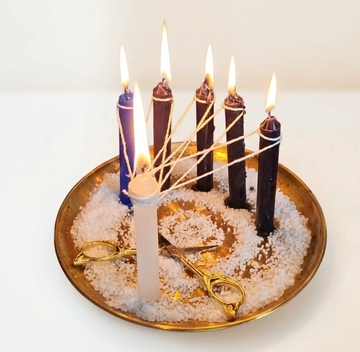Velas acesas ligadas por fios a uma vela branca, sobre um prato dourado com grãos brancos e uma tesoura dourada.