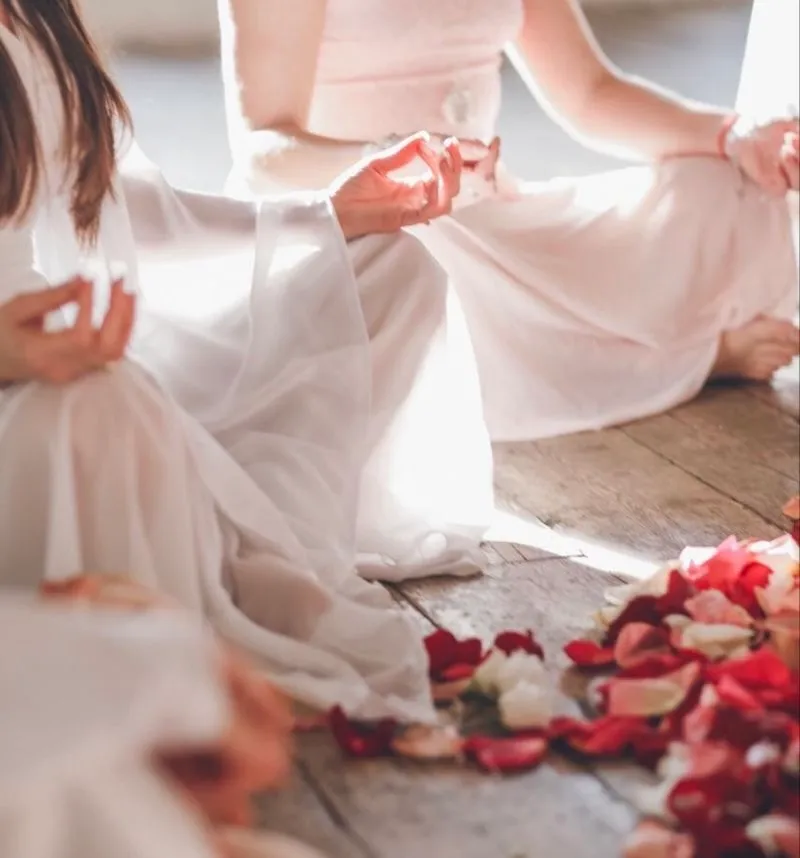 Mulheres de roupa clara sentadas no chão de madeira, em meditação, com pétalas de rosa ao redor.